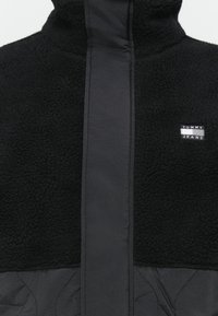 Zwarte fleecejas met een glad voorpaneel, een gestructureerd lichaam en een klein rechthoekig "Tommy Jeans" logo op de borst.