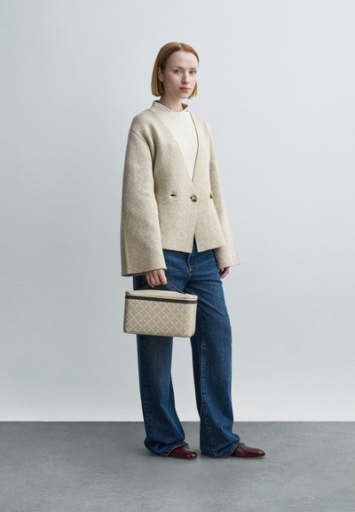 Μπεζ παλτό από μαλλί διπλής σειράς με φαρδιές μανσέτες, συνδυασμένο με εκρού τοπ και μπλε φαρδιά τζιν. Κρατάει μία μπεζ τσάντα με σχέδια.