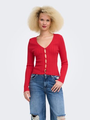 Kvinde med krøllet blondt hår iført en rød knappet ribbet cardigan og revnede blå jeans, stående med den ene hånd i lommen.