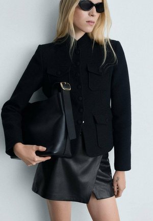 Black textured jacket with front pockets, paired with a black leather mini skirt. Model holds a black bag with a gold accent strap.