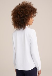 Vrouw met krullend haar die een langzijdig wit shirt en donkere broek draagt, met haar gezicht naar rechts tegen een effen beige achtergrond.