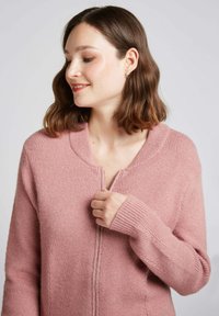 Cardigan zippé rose avec texture côtelée et col rond. Il présente des manches longues et une coupe légèrement ample, mettant en avant un matériau doux et chaud.