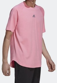 Růžové sportovní tričko s krátkým rukávem, kulatým výstřihem, raglánovými rukávy a malým tmavým logem na hrudi. Hladká textura, volný střih.