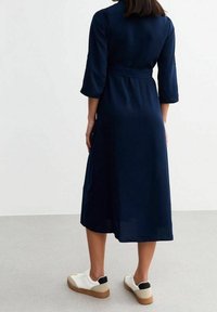 Marinblå midi-klänning med 3/4-ärmar och ett bälte i midjan, tillsammans med vita sneakers med svarta Accenter och en beige sula.