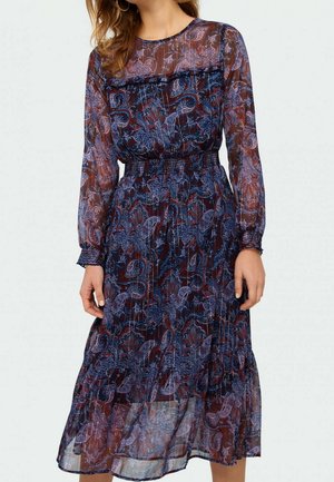 Frau trägt ein durchsichtiges Midi-Kleid mit langen Ärmeln, blauem und bordeauxfarbenem Paisley-Muster sowie geraffter Taille und Manschetten.