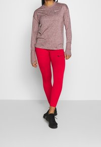 Top atletico bordeaux a manica lunga con scollo tondo, abbinato a leggings aderenti rossi con logo nero, e scarpe da corsa nere.