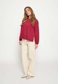 Tumepunane pika varrukaga pullover-sviiter, kombineeritud helevalgete laiade pükstega. Modell kannab teravate varvastega beeže kontsakingi. Lihtne, struktureeritud disain.