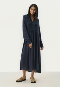 Navyblaues Langarmkleid mit Kragen, das vertikale Falten und eine geraffte Taille aufweist. Kombiniert mit beigen Sandalen mit offener Zehenpartie.