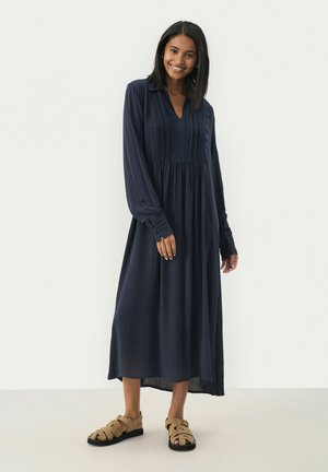 Mörkblå klänning med lång ärm och krage, med vertikala veck och resår i midjan. Matchas med tanfärgade sandals med öppen tå.