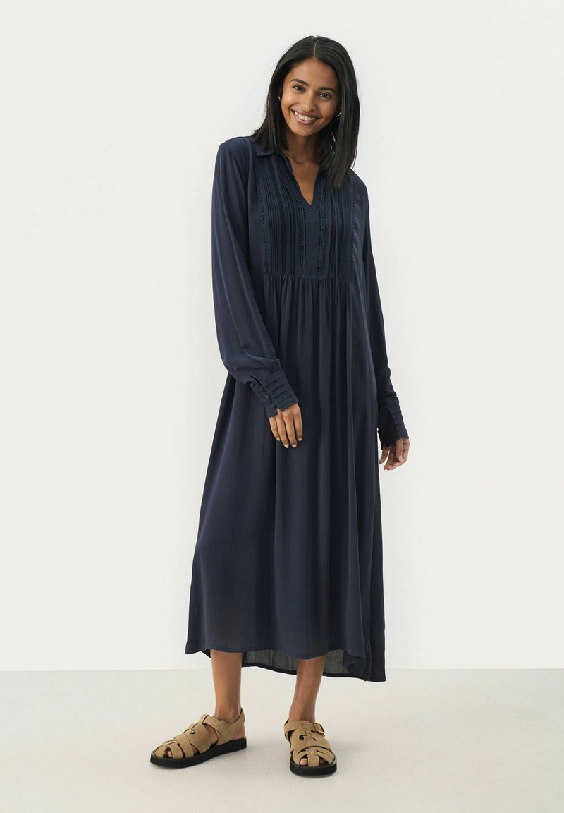 Navyblaues Langarmkleid mit Kragen, das vertikale Falten und eine geraffte Taille aufweist. Kombiniert mit beigen Sandalen mit offener Zehenpartie.