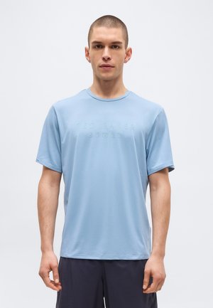 Νεαρός με κοντά μαλλιά φοράει ανοιχτό μπλε T-shirt Ted Baker Sport και σκούρα σορτς, στέκεται και κοιτάζει προς τα εμπρός πάνω σε απλό φόντο.