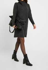 Vestito grigio lavorato a maglia con collo alto, abbinato a una borsa nera oversize e stivali alla caviglia. Le calze aggiungono texture e una silhouette elegante.