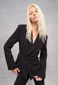 Blazer nero su misura con un design asimmetrico, caratterizzato da maniche lunghe e scollatura a V profonda. Tessuto liscio con una vestibilità strutturata.