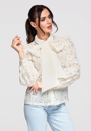 Femme aux cheveux foncés attachés en queue de cheval, portant un chemisier blanc en dentelle avec un grand nœud, associé à un jean bleu clair, posant devant un fond uni.