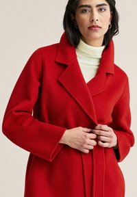 Punainen villakangastakki notkotulla kauluksella ja leveillä hihoilla, jossa on solmittava vyö vyötäröllä. Päällä on valkopohjainen ribbattu pooloneule.