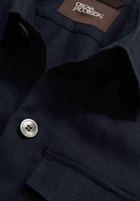 Navy blue button-up shirt made of textured fabric, featuring a silver button, a chest pocket, and a branded tag at the collar.