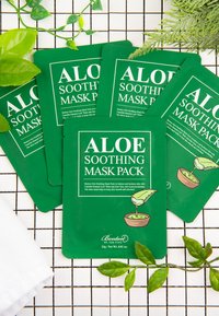 Pacchetti di maschere facciali verdi con design all'aloe caratterizzati da testo bianco. Circondati da piante verdi e un tessuto bianco su uno sfondo a griglia.