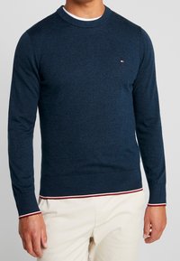 Suéter de punto azul marino con mangas largas, cuello redondo, puños y dobladillo acanalados con rayas rojas y blancas. Pequeño logotipo en el pecho.