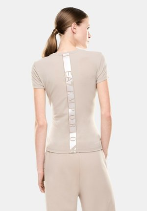 Donna con coda di cavallo castano chiaro che indossa un top a maniche corte beige e pantaloni, con una striscia riflettente verticale sulla schiena con la scritta "EA7 EMPORIO".