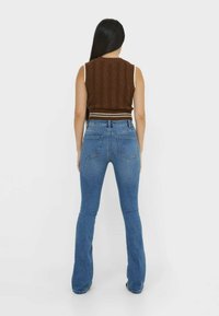 Hnědý bezrukávový top s kabelovým vzorem a žebrovaným lemem v kombinaci s modrými rozšířenými džíny. Oblečení má jednoduchý, přiléhavý střih.