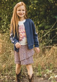 Marineblauw jack van een katoenmix met witte strepen aan de zijkant, licht t-shirt met print en rok met bloemen en ruches, gecombineerd met bruine cowboylaarzen.