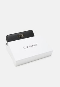 Black leather wallet with gold "CK" logo, partially visible in a white box featuring "Calvin Klein" printed in modern font.