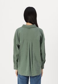 Blouse verte texturée avec un col, des manches longues et des détails de boutons aux poignets. Présente des plis au milieu du dos pour plus de volume.