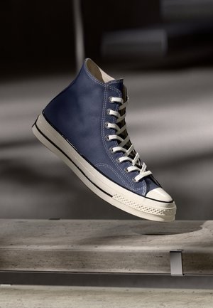 High-Top Sneaker in Marineblau mit weißen Schnürsenkeln und Gummisohle, der über einer Betonfläche vor dunklem Hintergrund schwebt.