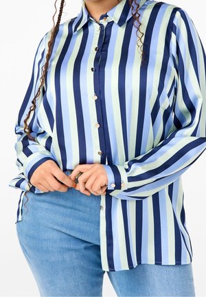 Person iført en langærmet skjorte med lodrette navy, lyseblå og bleggrønne striber og lyseblå jeans, hænder justerer skjortesømmen.