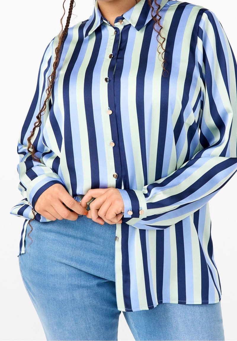 Personne portant une chemise à manches longues boutonnée avec des rayures verticales bleu marine, bleu clair et vert pâle, et un jean bleu clair, les mains ajustant l'ourlet de la chemise.