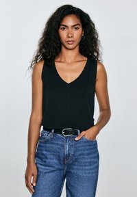 Joven con cabello rizado y negro, vistiendo una blusa negra sin mangas con escote en V, jeans azules con cinturón negro y pendientes de aro, de pie frente a un fondo gris.