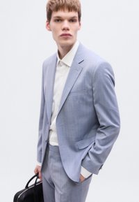 Ανοιχτό μπλε κοστούμι με κεντρικό πέτο και μία τσέπη. Λευκό πουκάμισο με κουμπιά από κάτω. Μαύρη τσάντα χειρός κρατημένη σε ένα χέρι.