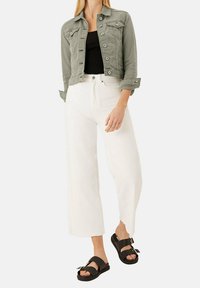 Veste en denim vert olive avec des boutons argentés, débardeur noir, pantalons blancs à jambes larges et sandales noires avec des accents à double lanière.