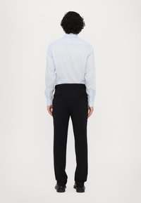 Lichtblauw langemouwen overhemd en zwarte op maat gemaakte broek. Het overhemd heeft een soepele textuur; de broek heeft een traditionele pasvorm en discrete zakken.