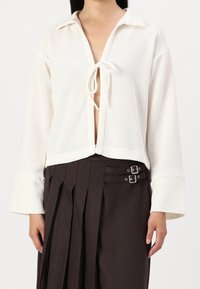 Žena v bílém pleteném topu s dlouhým rukávem a předníma zavazovacíma provázkama, doplněném o tmavě hnědou plisovanou sukni se dvěma stříbrnými sponami.