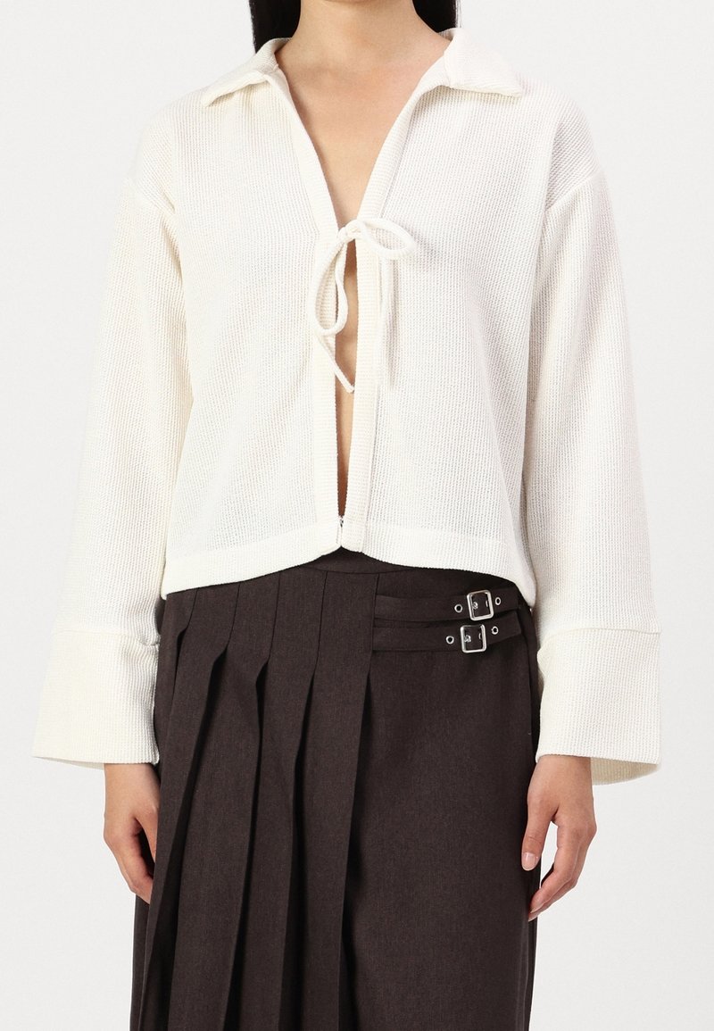Žena v bílém pleteném topu s dlouhým rukávem a předníma zavazovacíma provázkama, doplněném o tmavě hnědou plisovanou sukni se dvěma stříbrnými sponami.