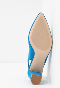 Niebieski zamszowy but na wysokim obcasie, z wydłużonym noskiem i wycięciami po bokach, posiadający teksturowaną beżową gumową podeszwę dla lepszej przyczepności.