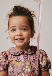Vestido de algodón con un estampado floral en tonos rosa-marrón, mangas cortas abullonadas y cuello redondo. Sujetado con un lazo, el niño sonríe, mostrando ojos brillantes.