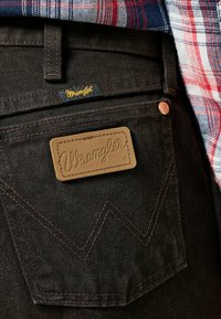 Jean en denim sombre avec un patch logo Wrangler, des accessoires en cuivre et une couture en zigzag distinctive sur la poche arrière.