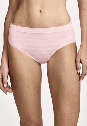 Frau trägt hellrosa nahtlose Unterwäsche, steht mit entspannten Armen an den Seiten vor einem einfarbigen Hintergrund.