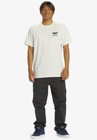 White cotton t-shirt featuring a butterfly graphic and "Quiksilver" text on the left chest, paired with black pants and blue sneakers.