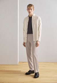 Camicia button-up beige chiaro, t-shirt blu navy sotto, pantaloni grigio chiaro e scarpe nere. Tessuto liscio, design sartoriale, accenti minimi.