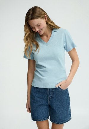 Kvinde med langt hår iført en lyseblå kortærmet V-hals top og mørke denimshorts, smilende og kiggede ned med den ene hånd i lommen.