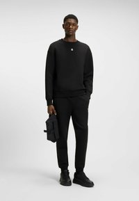 Black sweatshirt with a subtle logo on the chest, paired with black sweatpants. The model holds a black bag with a buckle.
