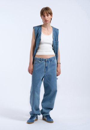 Denim vest, white fitted tank, blue oversized jeans, and blue sneakers with three white stripes. Notable relaxed fit and minimal hardware.