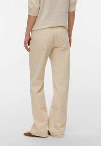Personne portant un pantalon beige à jambes larges et un pull beige clair, debout avec les mains détendues le long du corps, devant un fond uni.