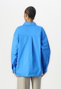 Camicia oversize azzurro chiaro in cotone con colletto aperto, spalle scese e polsini con bottone. Sul retro presenta un dettaglio di cucitura orizzontale.