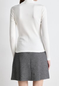 Vista posteriore di una persona che indossa un top a maniche lunghe bianco a coste e una gonna a scacchi bianco e nero, in piedi su uno sfondo semplice.