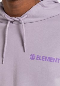 Primo piano di una persona che indossa un hoodie color lilla chiaro con spesse corde di regolazione e un logo parziale "ELEMENT" in viola sul petto.