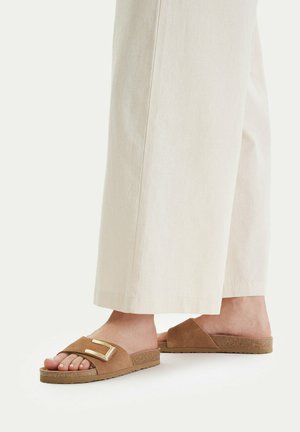 Pies calzando sandalias planas de ante color marrón claro con detalle de hebilla dorada, combinadas con pantalones anchos y sueltos de tono beige claro sobre un fondo liso.